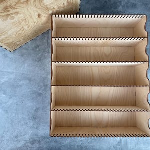 May include: A light-colored wooden organizer with five shelves and a matching lid. The organizer has a rectangular shape with curved edges and is made of thin wooden panels. The lid features a detailed tree design.