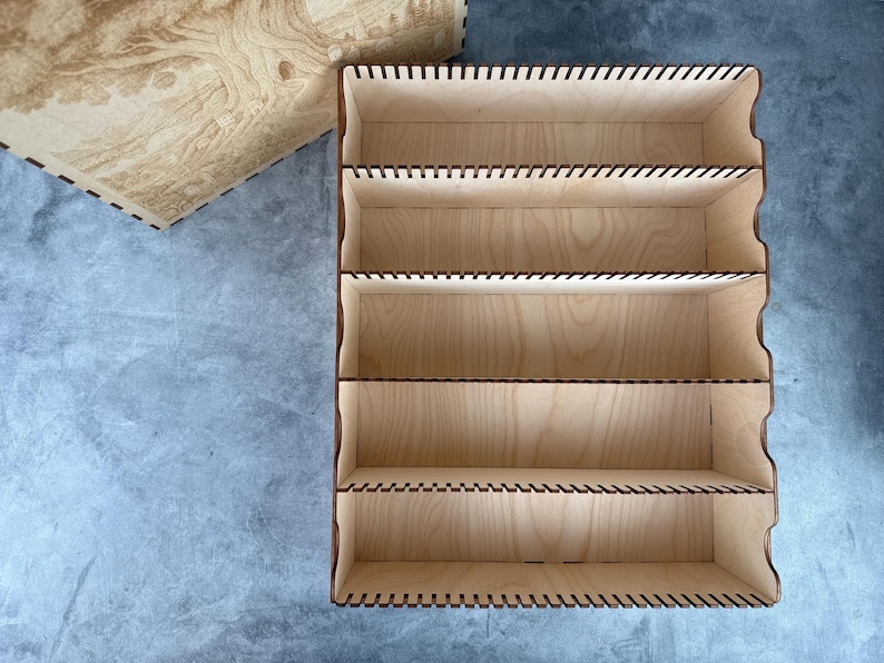 May include: A light-colored wooden organizer with five shelves and a matching lid. The organizer has a rectangular shape with curved edges and is made of thin wooden panels. The lid features a detailed tree design.
