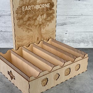 May include: A wooden organizer with the text "EARTHBORNE RANGERS" and a tree design. The organizer has multiple compartments and decorative cutouts. The lid is open, revealing the interior.