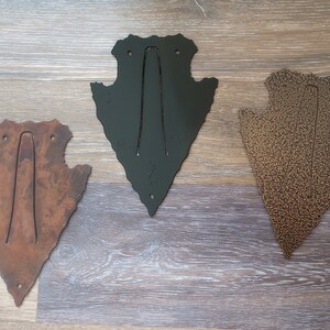 May include: Three metal arrowhead-shaped wall hooks in different finishes: rusty brown, black, and copper. Each hook has a simple, curved design for hanging items.