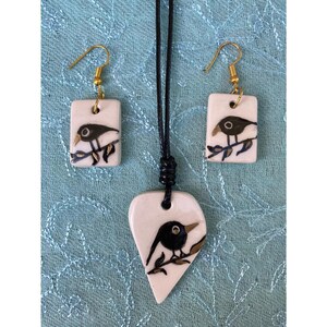 May include: A white ceramic necklace and earrings set with black bird illustrations. The necklace is a heart shape and the earrings are rectangular.