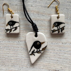 May include: A black ceramic necklace and earrings set with a heart-shaped pendant and square earrings. Each piece features a black bird perched on a gold branch.