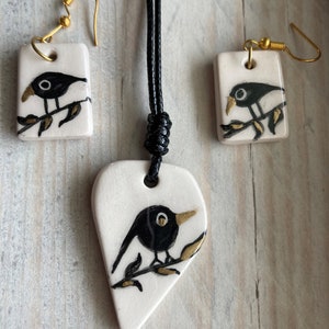 May include: A black bird on a branch necklace and earrings set. The necklace is a heart-shaped pendant with a black bird on a branch. The earrings are square-shaped with a black bird on a branch. The jewelry is made of white ceramic with gold accents.