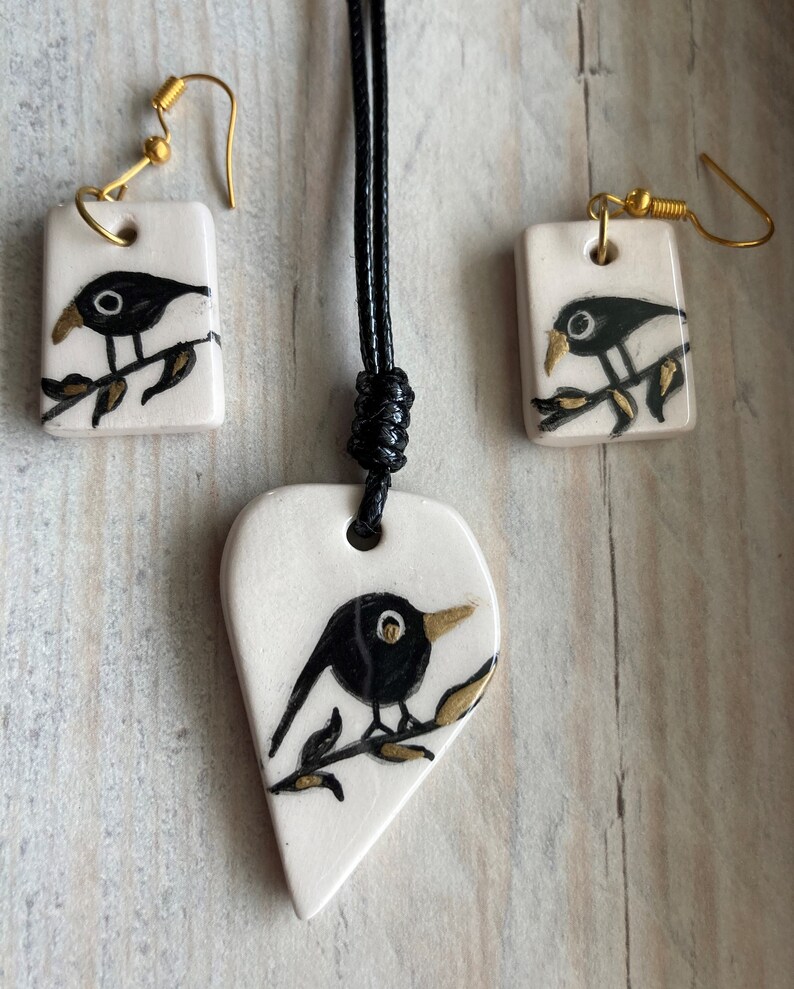 May include: A black bird on a branch necklace and earrings set. The necklace is a heart-shaped pendant with a black bird on a branch. The earrings are square-shaped with a black bird on a branch. The jewelry is made of white ceramic with gold accents.