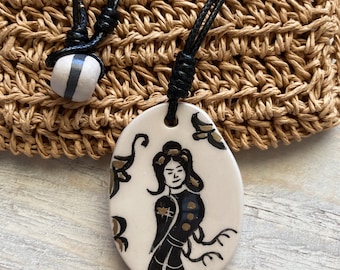 Pode incluir: Um pendente de cerâmica com uma ilustração pintada à mão de uma figura em preto e dourado sobre uma base oval branca. O pendente está suspenso num cordão preto com um nó ajustável e uma pequena conta branca com uma risca cinzenta.
