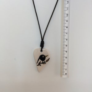 May include: A black and white ceramic pendant necklace featuring a bird perched on a branch. The pendant is shaped like a heart and is attached to a black cord.
