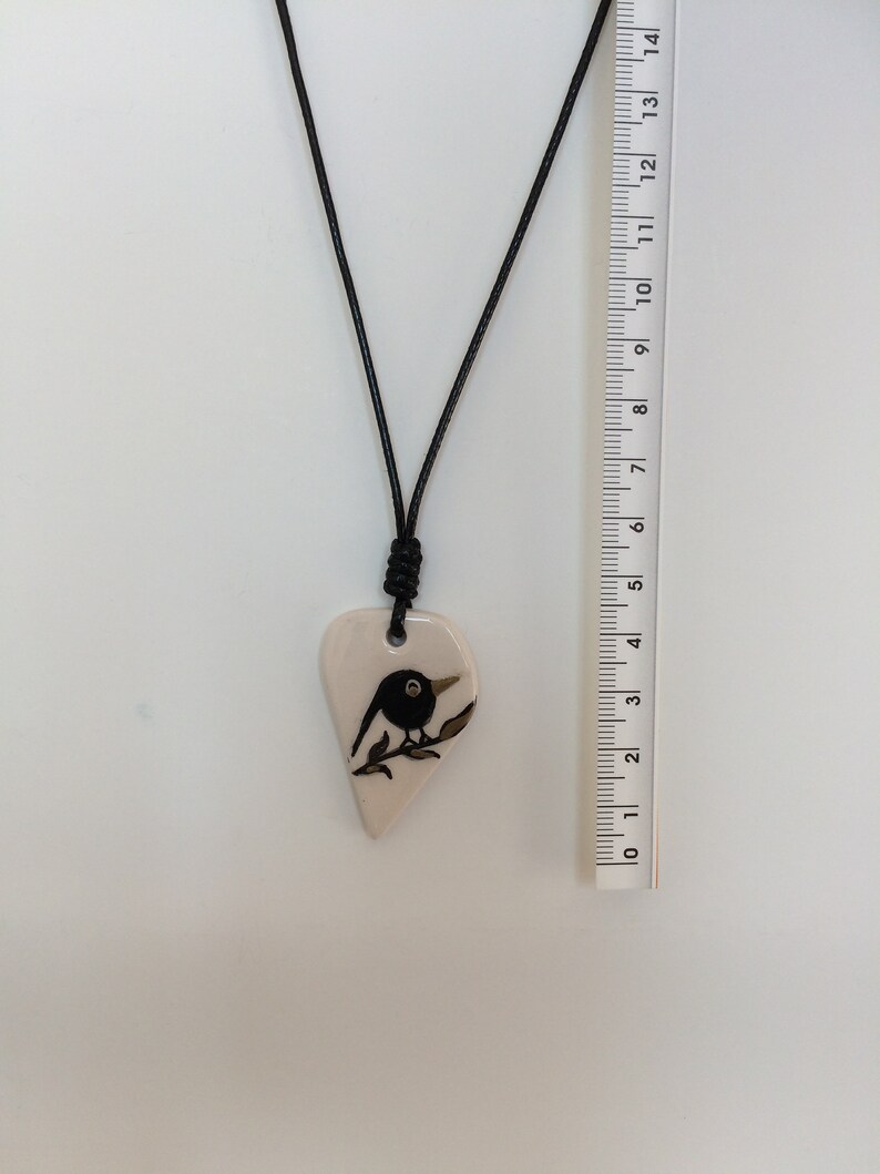 May include: A black and white ceramic pendant necklace featuring a bird perched on a branch. The pendant is shaped like a heart and is attached to a black cord.