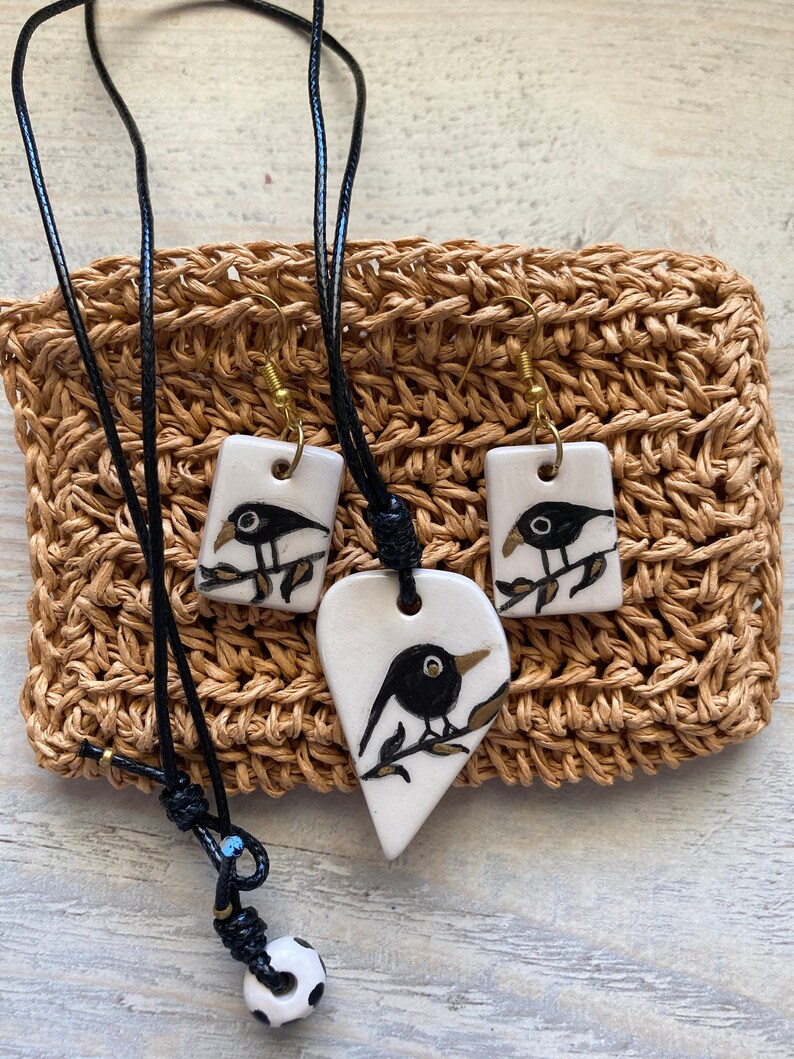 May include: A set of black bird ceramic jewelry, including a necklace and earrings. The necklace features a heart-shaped pendant with a black bird on a branch, while the earrings are rectangular with black birds on branches. The jewelry is displayed on a woven brown background.