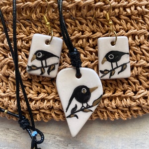 May include: A set of black and white ceramic bird pendant necklaces with black leather cords. The pendant necklaces feature a small black bird on a branch with gold accents. The set also includes a pair of matching earrings.