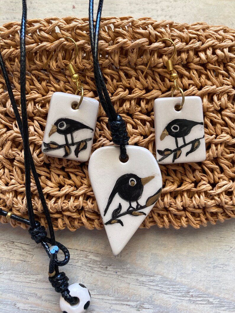 May include: A set of black and white ceramic bird pendant necklaces with black leather cords. The pendant necklaces feature a small black bird on a branch with gold accents. The set also includes a pair of matching earrings.