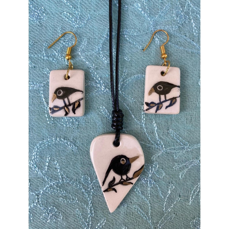 May include: A white ceramic necklace and earrings set with black bird illustrations. The necklace is a heart shape and the earrings are rectangular.