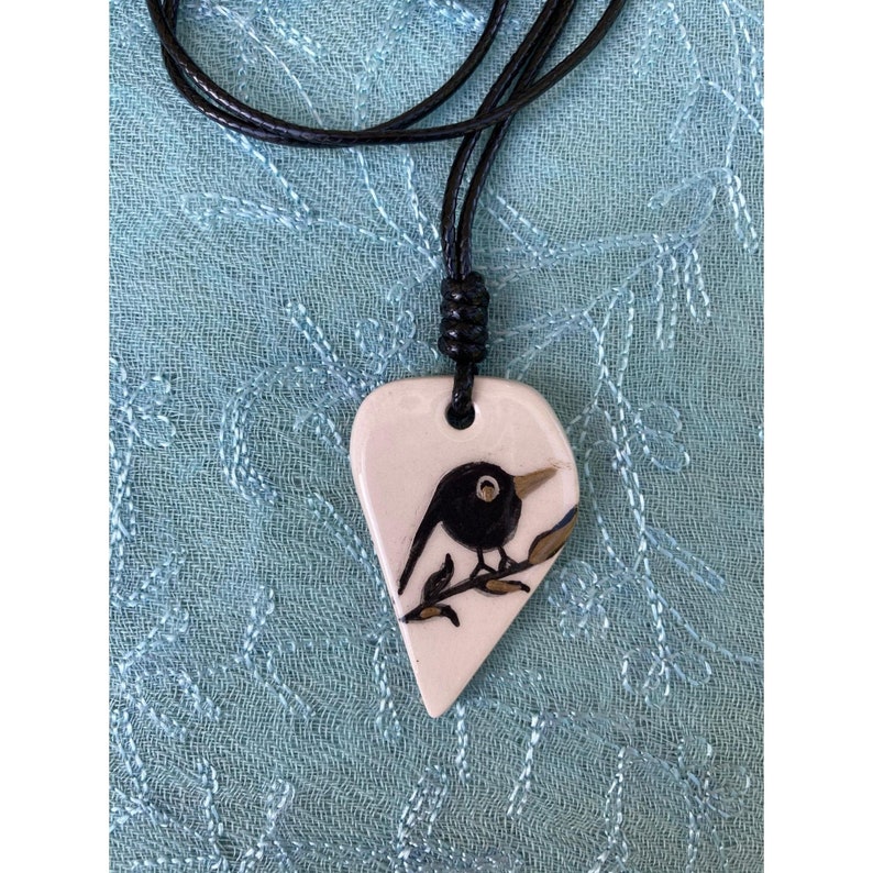 May include: A white ceramic heart-shaped pendant necklace with a black bird on a branch, the bird has a gold eye and the branch has gold accents. The necklace has a black cord.