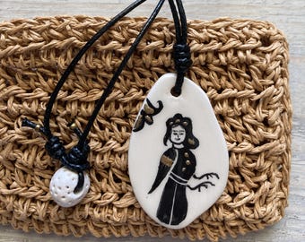 Pode incluir: Um pingente de cerâmica branca com uma ilustração pintada à mão em preto e dourado de uma figura, suspenso por um cordão preto. Uma conta de cerâmica branca menor e esférica com várias reentrâncias está presa ao cordão perto do pingente.