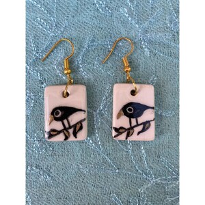 May include: A pair of ceramic earrings with a black bird design on a white background. The earrings have gold hooks.