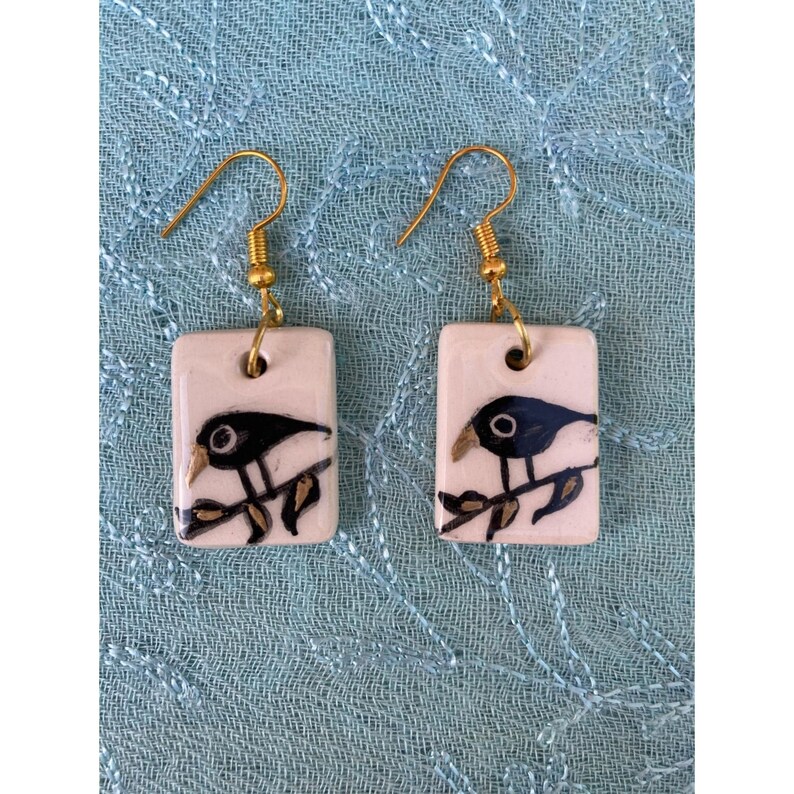May include: A pair of ceramic earrings with a black bird design on a white background. The earrings have gold hooks.