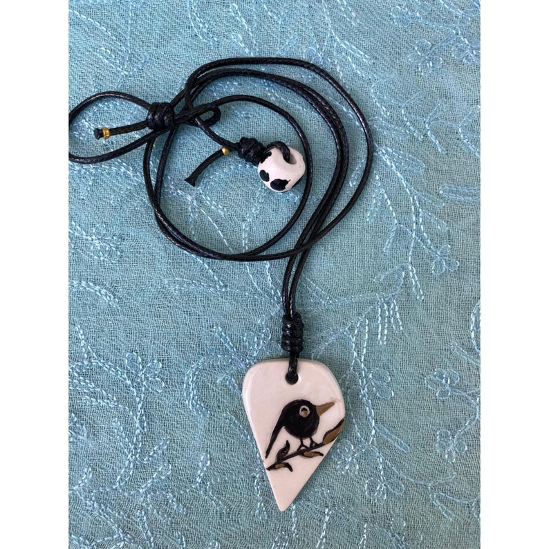 May include: A black cord necklace with a white ceramic pendant shaped like a heart. The pendant features a black bird perched on a branch with gold accents.