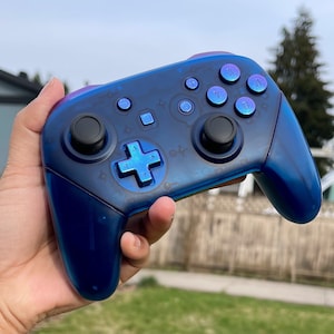 以下が含まれることがあります： テクスチャ加工が施された青と紫のNintendo Switchコントローラー。コントローラーには、方向パッド、4つのフェイスボタン、2つのアナログスティック、2つのショルダーボタンがあります。