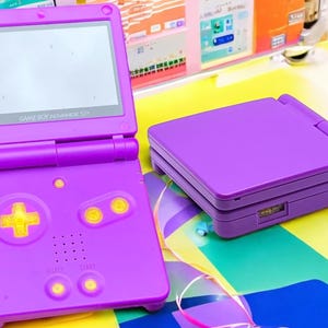 May include: A purple Game Boy Advance SP handheld video game console with a white screen and yellow buttons. The console is closed and sitting on a colourful surface.