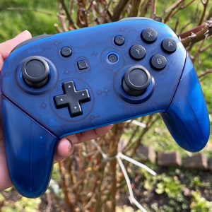 Puede incluir: Un mando Nintendo Switch transparente azul con botones negros y una palanca de mando negra. El mando tiene un diseño transparente, lo que permite ver los componentes internos.