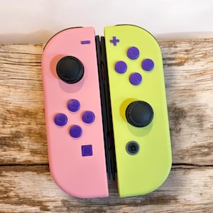 May include: A pair of Nintendo Switch Joy-Con controllers. One is pink, the other is lime green. Each controller has black joysticks and purple buttons. The controllers are resting on a wooden surface.