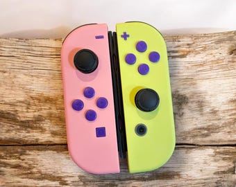 May include: A pair of Nintendo Switch Joy-Con controllers. One is pink, the other is lime green. Each controller has black joysticks and purple buttons. The controllers are resting on a wooden surface.