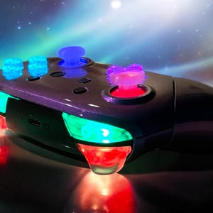 May include: A black and purple wireless games controller with illuminated blue, green, and red lights. The controller has multiple buttons and joysticks, with a sleek, ergonomic design. The background features a starry, galaxy-themed design.