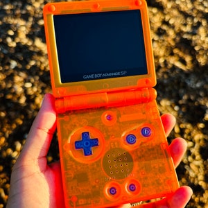 May include: A translucent orange Game Boy Advance SP handheld gaming console with a black screen and blue buttons. The console is held in a hand.