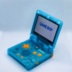 K&ouml;nnte beinhalten: Ein durchsichtiger blauer Game Boy Advance SP mit gelben Kn&ouml;pfen. Der Bildschirm ist ge&ouml;ffnet und zeigt das Game Boy-Logo.