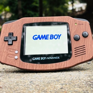 Puede incluir: Una consola de juegos portátil Game Boy Advance de madera marrón con una pantalla negra que muestra el texto "GAME BOY" en azul. La consola tiene botones negros y una almohadilla direccional negra.