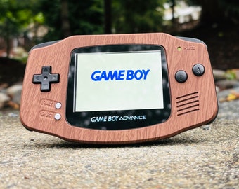 May include: A brown wood-grain Game Boy Advance handheld gaming console with a black screen displaying the text "GAME BOY" in blue. The console has black buttons and a black directional pad.