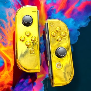 Puede incluir: Dos mandos Joy-Con dorados para Nintendo Switch con botones negros sobre un fondo colorido.
