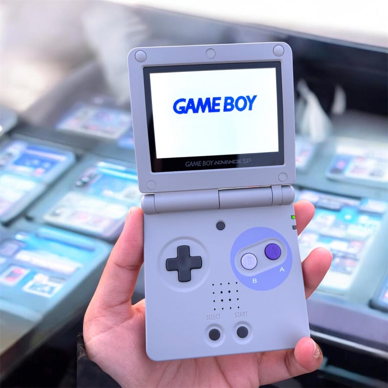 May include: A light gray Game Boy Advance SP handheld game console. The screen displays "GAME BOY" in blue. The console has a directional pad, A and B buttons, and start/select buttons. The text "GAME BOY ADVANCE SP" is below the screen.