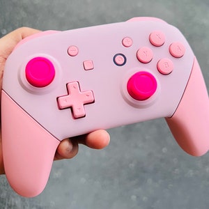 May include: A pink Nintendo Switch controller with pink buttons. The controller has a pink and white design.