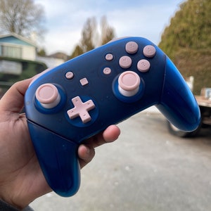 May include: A blue and pink video game controller. The controller has a dark blue body with pink buttons, including the directional pad, face buttons, and joysticks. The controller is held in a hand, with a blurred outdoor background.