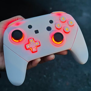 May include: A white Nintendo Switch controller with red and yellow illuminated buttons. The controller has a white, textured surface and a black faceplate.