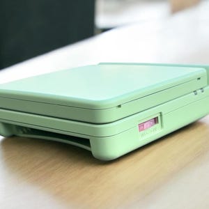 May include: A light green handheld gaming console with a pink power button and a sliding switch labeled "ON - OFF".