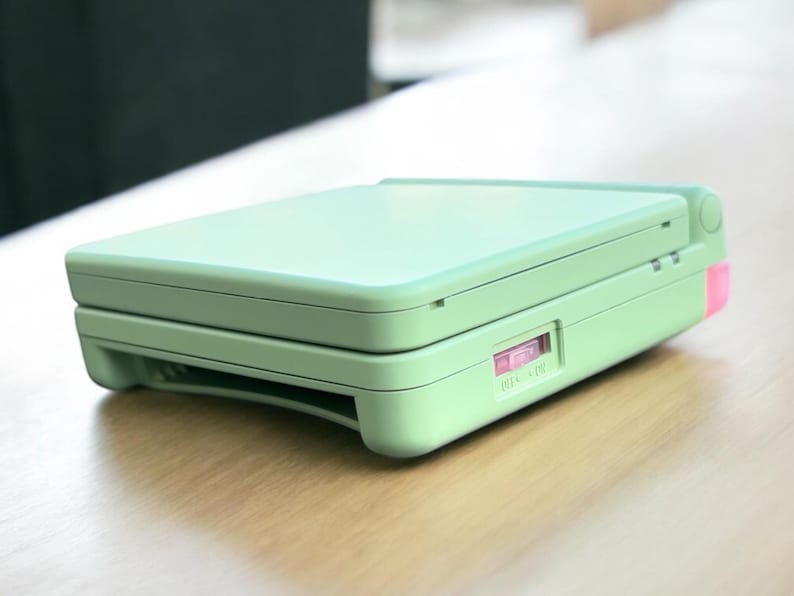 May include: A light green handheld gaming console with a pink power button and a sliding switch labeled "ON - OFF".