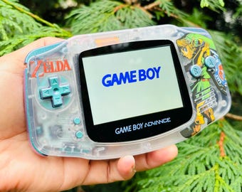 May include: A clear, translucent Game Boy Advance handheld gaming console with a blue and green Zelda themed design. The screen displays the words "GAME BOY" in blue text. The console has a blue and green button pad and a blue and green "SELECT" button.