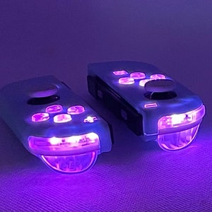 Może przedstawiać: Dwa białe bezprzewodowe kontrolery do gier z fioletowymi diodami LED. Kontrolery mają różne przyciski i joystick.