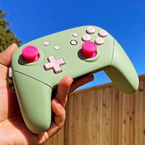 Puede incluir: Un mando de Nintendo Switch verde y rosa con botones rosas. El mando está siendo sostenido en la mano de una persona.