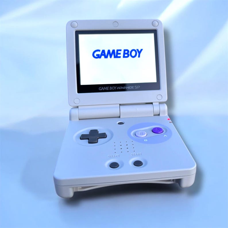 May include: A white Game Boy Advance SP handheld game console, open and displaying the word "GAME BOY" in blue on the screen. The console has a directional pad, buttons, and the text "GAME BOY ADVANCE SP".