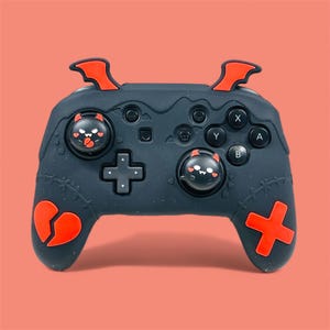 May include: Black gaming controller with red detailing, including horns, a broken heart, and a plus sign. The joystick caps feature a devilish face design. Buttons are labelled X, Y, A, and B. The controller is set against a coral background.