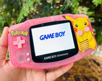 May include: A pink Game Boy Advance handheld video game console with a Pikachu design. The console has a black screen with the text "GAME BOY" displayed. The console also has a pink button and a pink directional pad.