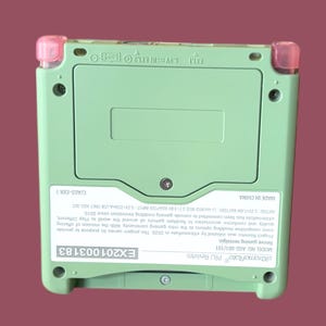 May include: A green Game Boy Advance SP with pink accents. The back of the device has a sticker with the text "eiktreme Rate PRJ Revivtro", "MODEL NO AGS-001/101", "Revive gaming nostalgia", "EX201003183", and a description of the project. The text "MADE IN CHINA" and "C/AGS-EXR 1" are also visible.