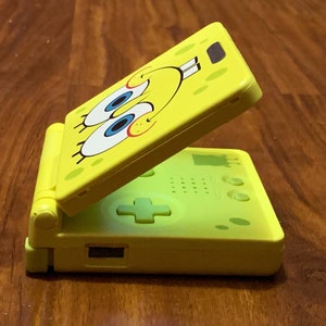 Puede incluir: Una consola de videojuegos portátil amarilla con un diseño de Bob Esponja en la solapa superior. La consola está abierta y la pantalla es visible.