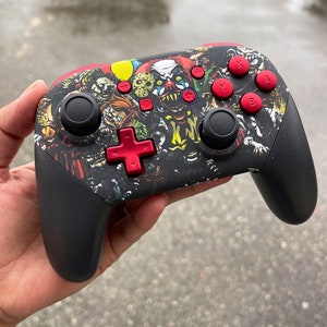 May include: A black Nintendo Switch controller with a custom skin featuring a horror movie theme. The skin features various horror movie characters, including a clown, a zombie, and a monster. The controller has red buttons and a red directional pad.