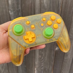 Può includere: Un controller Nintendo Switch giallo traslucido con pulsanti verdi. Il controller ha un design trasparente, quasi traslucido, che consente di vedere i componenti interni.