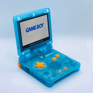 K&ouml;nnte beinhalten: Eine durchsichtige blaue Game Boy Advance SP Handheld-Spielkonsole mit einem gelben Knopf und dem Text "GAME BOY" auf dem Bildschirm.