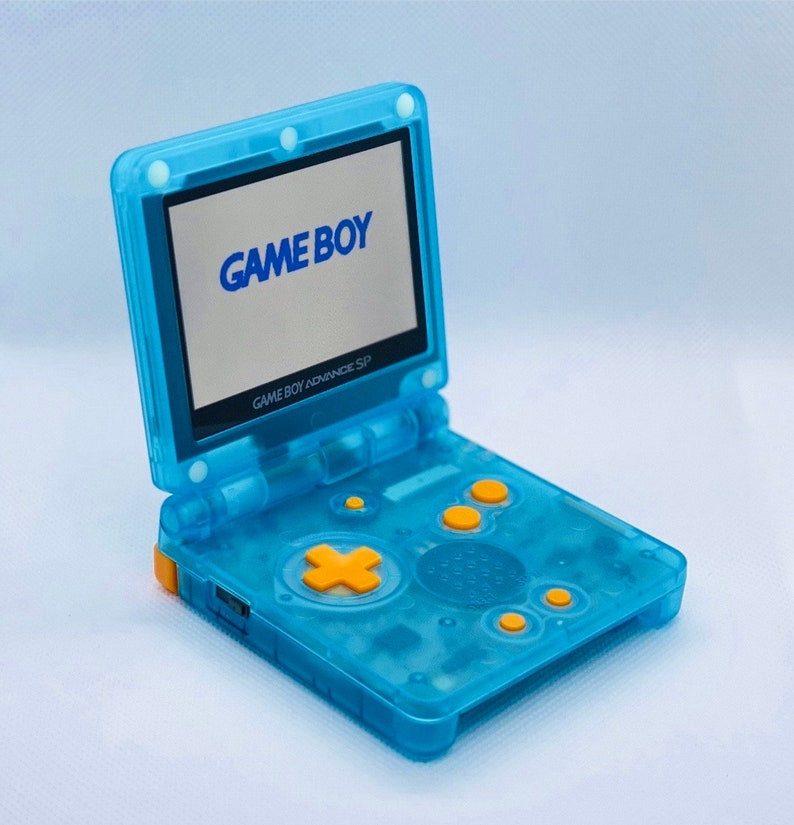 K&ouml;nnte beinhalten: Eine durchsichtige blaue Game Boy Advance SP Handheld-Spielkonsole mit einem gelben Knopf und dem Text "GAME BOY" auf dem Bildschirm.