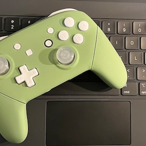 May include: A green and white video game controller with white buttons and a white plus sign on a black laptop keyboard.
